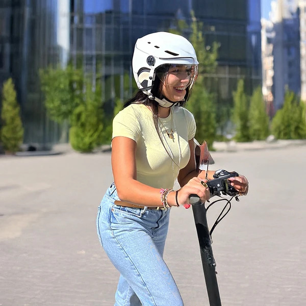 CASQUE URBAN MOOV VISOR 10 Femme portant le casque Urban Moov Visor blanc avec visière sur une trottinette électrique en ville