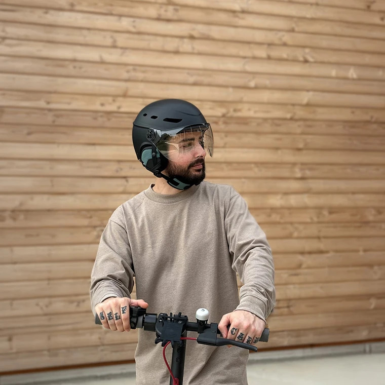 CASQUE URBAN MOOV VISOR 11 Homme portant le casque Urban Moov Visor noir avec visière relevée sur une trottinette électrique